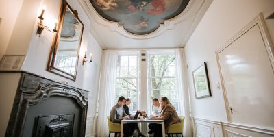 Professionals in overleg in een historische stijlkamer van EBC Amsterdam met een indrukwekkend plafondschilderij en klassieke schouw.